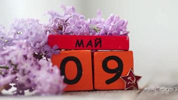 Wooden calendar with russian text May 9th. Victory Day. Lilac petals fall on wooden background video