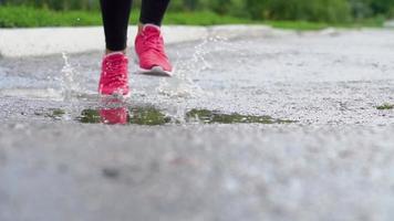 Close up of legs of a runner in sneakers. Sports woman jogging outdoors, stepping into muddy puddle. Single runner running in rain, making splash. Slow motion video