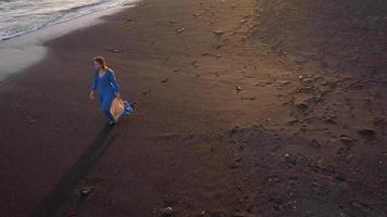 parte superior ver de un niña en un azul vestir y sombrero caminando en el playa con negro arena, espumoso olas de el atlántico océano. tenerife, canario islas, España video