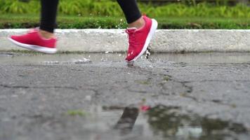 stänga upp av ben av en löpare i sneakers. sporter kvinna joggning utomhus, stepping in i grumlig pöl. enda löpare löpning i regn, framställning stänk video