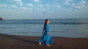 mujer en hermosa azul vestir corriendo a lo largo un negro volcánico playa video