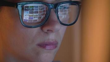 Woman in glasses looking on the monitor and surfing Internet at night. The monitor screen is reflected in the glasses video