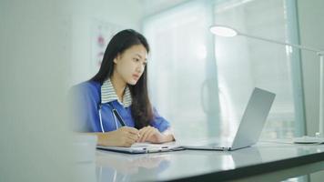 Young female doctor in white coat working on laptop in modern hospital office room, therapist typing on computer consult patient online, makes research, noting, takes useful information from internet video