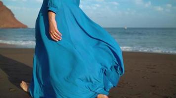 piernas de un mujer en hermosa azul vestir caminando a lo largo un negro volcánico playa video
