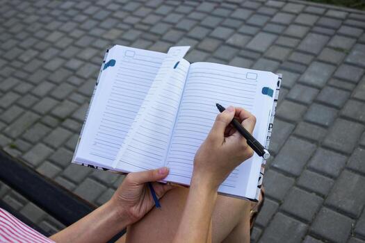 Top view image of open pad in female hands with pen sitting on stairs in the park. Blank sheets of small notebook overhead view photo
