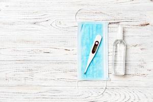 Top view of surgical mask, digital thermometer and alcohol hand sanitizer on wooden background. Health care concept with copy space photo