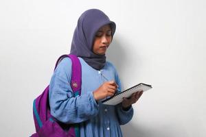 Young Asian girl student in hijab writing something on the book isolated on white background. photo