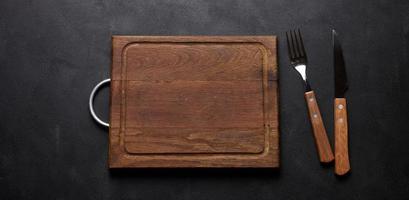 Fork and knife with wooden handle and cutting board on black background, top view photo