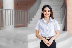 Beautiful Asian young woman student in Thai student uniform is stand under the school building smiling and looking at camera in university background. photo