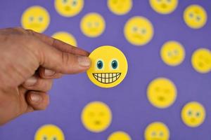 Closeup shot of hand holding yellow smiley face emoji between sad emojis on purple background photo