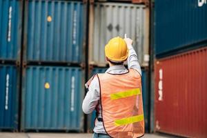Asian technician dock worker in protective safety jumpsuit uniform and with hardhat and use laptop computer at cargo container shipping warehouse. transportation import,export logistic industrial photo