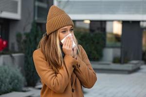 ill beautiful young woman sneezing and blowing nose in napkin. Sick businesswoman in business style sneeze at street. Unhealthy employee. Outdoor. Virus symptoms. Cold disease. photo