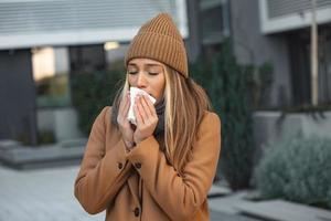 ill beautiful young woman sneezing and blowing nose in napkin. Sick businesswoman in business style sneeze at street. Unhealthy employee. Outdoor. Virus symptoms. Cold disease. photo