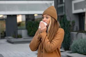 ill beautiful young woman sneezing and blowing nose in napkin. Sick businesswoman in business style sneeze at street. Unhealthy employee. Outdoor. Virus symptoms. Cold disease. photo