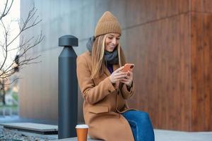 Image of beautiful stylish woman sitting on street bench with legs crossed on sunny day and holding mobile phone photo