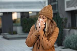ill beautiful young woman sneezing and blowing nose in napkin. Sick businesswoman in business style sneeze at street. Unhealthy employee. Outdoor. Virus symptoms. Cold disease. photo