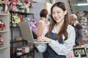 Portrait of one young Asian female florist worker in apron with laptop in bright and beautiful flower shop store, smiling and looking at camera, small business occupation, happy SME entrepreneur. photo