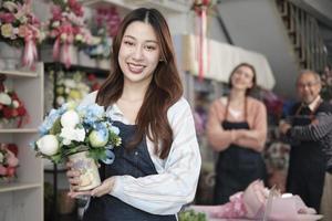 Beautiful young Asian female florist worker with vase of blossom smiles and looks at camera in front of colleagues team in colorful flower shop, small business occupation, happy SME entrepreneur. photo