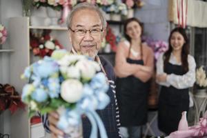 Senior Asian male florist owner in apron gives bunch of blossom smiles and looks at camera in front of her colleague's team in colorful flower shop, small business occupation, happy SME entrepreneur. photo