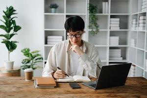 Male student taking notes from a book at library, Young asian sitting at desk doing assignments in college library photo