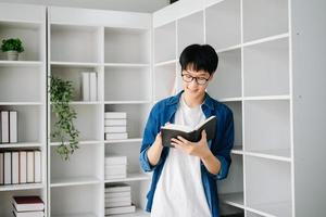 masculino estudiante tomando notas desde un libro a biblioteca, joven asiático sentado a escritorio haciendo asignaciones en Universidad biblioteca foto