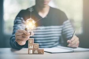 Businessman hand holding light bulb with icon on wood cube and working on desk, Creativity, and innovation are keys to success. Concept of new idea and innovation with Brain and light bulbs. photo