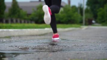 stänga upp av ben av en löpare i sneakers. sporter kvinna joggning utomhus, stepping in i grumlig pöl. enda löpare löpning i regn, framställning stänk video