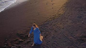 Top view of a girl in a blue dress and hat walking on the beach with black sand, foaming waves of the Atlantic Ocean. Tenerife, Canary Islands, Spain video