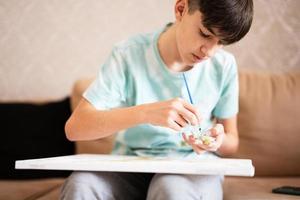 Young male teenager painting picture by numbers on canvas in living room at home. Hobby and leisure concept. photo