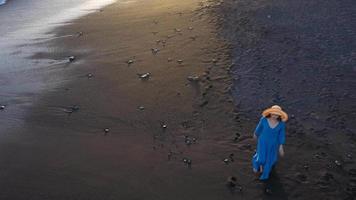 parte superior ver de un niña en un azul vestir y sombrero caminando en el playa con negro arena, espumoso olas de el atlántico océano. tenerife, canario islas, España video