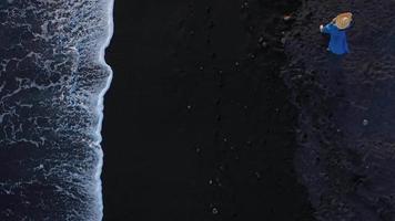 Top view of a girl in a blue dress and hat walking on the beach with black sand, foaming waves of the Atlantic Ocean. Tenerife, Canary Islands, Spain video