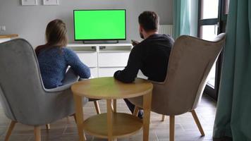 Man and woman are sitting in chairs, watching TV with a green screen, discuss what they saw and switching channels with video