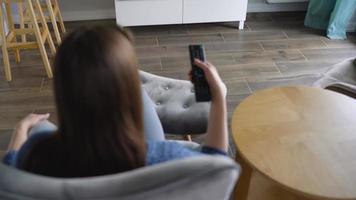 donna è seduta nel un' sedia, Guardando tv con un' verde schermo, commutazione canali con un' a distanza controllo. croma chiave video