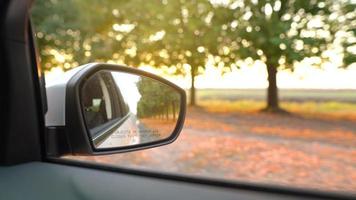 Visualizza a partire dal il retrovisore specchio come auto unità attraverso il strada circondato di alberi a tramonto video