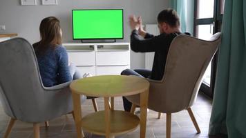 Man and woman are sitting in chairs, watching TV with a green screen, discuss what they saw and switching channels with video