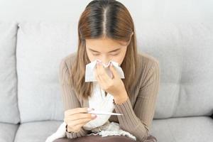 joven asiático mujer teniendo alto fiebre mientras comprobación cuerpo temperatura, hembra estornudos y líquido nariz con estacional influenza, alérgico, digital termómetro, virus, coronavirus, enfermedad, respiratorio foto