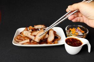 Right hand use chopsticks clamping grilled pork shoulder slice in white rectangle dish with hot and sour Thai style sauce in white cup and vegetable slice in vinegar glass cup on black background. photo