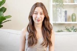 Smiling positive, attractive asian young woman wearing casual dress, portrait of beautiful brunette her with long brown hair, feeling happy looking to camera sitting on sofa, isolated on background. photo