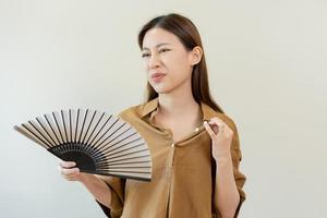 sufrimiento verano calor ataque, caliente clima, cansado asiático joven mujer, niña sudoroso y sediento, refrescante con mano en soplo, ola ventilador a ventilación cuando temperatura alto a hogar, casa en antecedentes foto