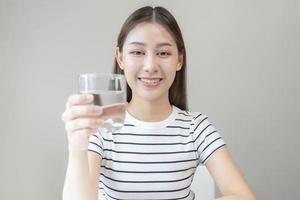 Happy beautiful, asian young woman, girl drinking, sip fresh glass of water for hydration of body, holding transparent glass in her hand, thirsty at home. Health care, healthy lifestyle concept. photo