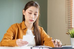 asiático joven empleado, negocio mujer calcular impuesto ingresos y gastos, facturas, crédito tarjeta para pago o día de paga en mesa a hogar oficina. financiero, Finanzas personas concepto. foto