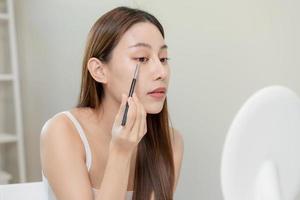 Happy beauty concept, cute asian young woman, girl smile, make up face by an eyeshadow brush on her eyes, looking into mirror in morning routine at home. People look with natural fashion style. photo