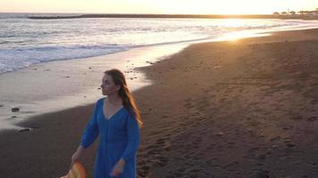 parte superior ver de un niña en un azul vestir y sombrero caminando en el playa con negro arena, espumoso olas de el atlántico océano. tenerife, canario islas, España video