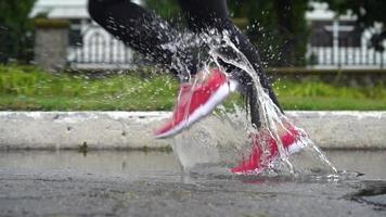 stänga upp av ben av en löpare i sneakers. sporter kvinna joggning utomhus, stepping in i grumlig pöl. enda löpare löpning i regn, framställning stänk video