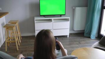 Frau ist Sitzung im ein Stuhl, Aufpassen Fernseher mit ein Grün Bildschirm, Schalten Kanäle mit ein Fernbedienung Kontrolle. Chroma Schlüssel video