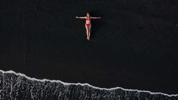 Haut vue de une fille dans une rouge maillot de bain mensonge sur une noir plage sur le le surf doubler. côte de le île de Ténérife, canari video