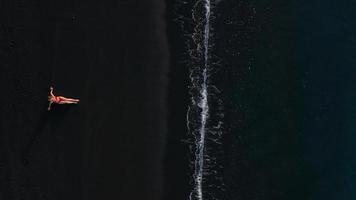 oben Aussicht von ein Mädchen im ein rot Badeanzug Lügen auf ein schwarz Strand auf das Surfen Linie. Küste von das Insel von Teneriffa, Kanarienvogel video