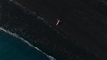 parte superior ver de un niña en un rojo traje de baño acostado en un negro playa en el navegar línea. costa de el isla de tenerife, canario video