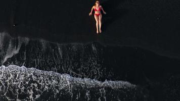 oben Aussicht von ein Mädchen im ein rot Badeanzug Lügen auf ein schwarz Strand auf das Surfen Linie. Küste von das Insel von Teneriffa, Kanarienvogel video