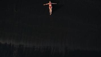 Haut vue de une fille dans une rouge maillot de bain mensonge sur une noir plage sur le le surf doubler. côte de le île de Ténérife, canari video